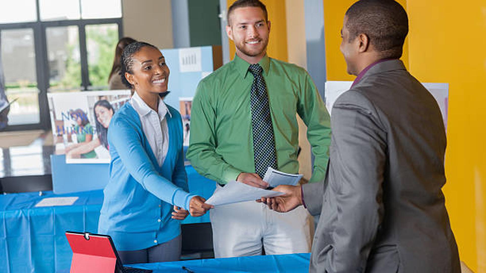 Two well dressed people talking to, and handing a piece of paper to a person in a suit over a table