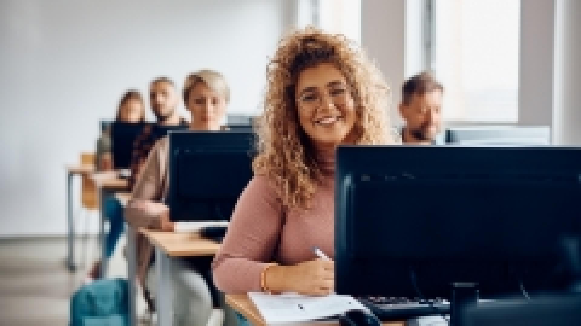 A classroom of people behind computer monitors in a row from front to back.