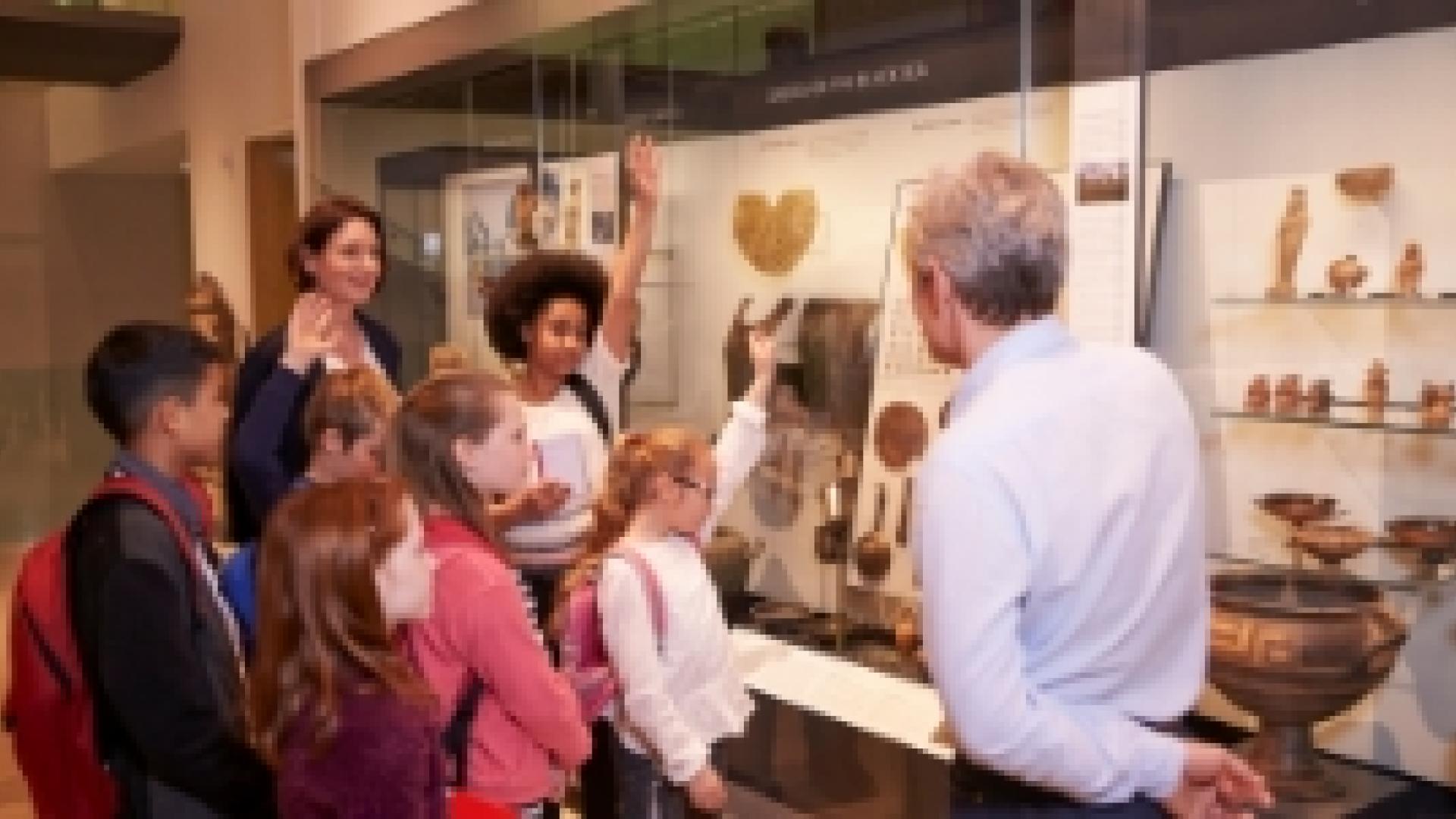 A museum tour led by a male presenting person, the school children on the tour have their hands up to ask questions